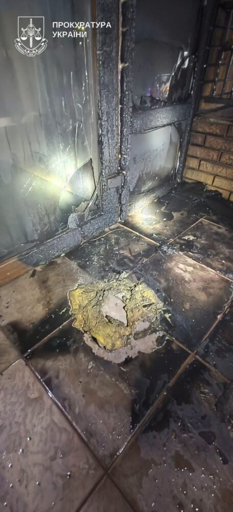 Charred doorway and scorched interior with melted metal and broken glass; damaged tiled floor with a large circular hole.