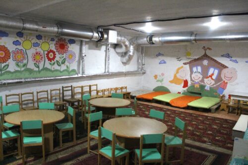 Playroom for kids with round tables, small green chairs, and bright wall murals of flowers and cartoon characters. A cushioned seating area sits to the side under an exposed ceiling.