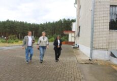 Three adults walking together on a brick path beside a beige brick building, with a forested area in the background.