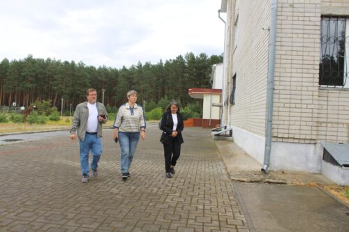 Three adults walking together on a brick path beside a beige brick building, with a forested area in the background.