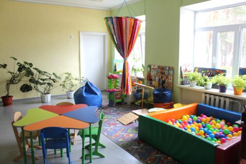 Bright children's playroom with colorful tables and chairs, a ball pit, bean bags, and plants by the window.