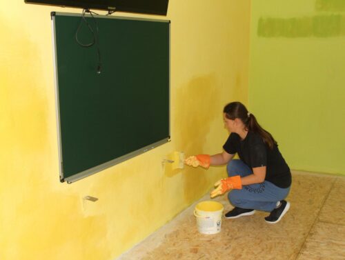 Woman kneeling by a yellow wall, rollers applying paint with orange gloves and a bucket of paint nearby beside a green chalkboard.