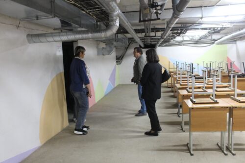 Three people stand in a basement classroom hallway with desks and chairs stacked on top, exposing ductwork above and a geometric mural on the wall.