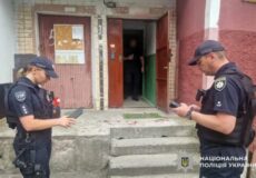 Two Ukrainian police officers in dark uniforms and caps stand on concrete steps outside a building, looking at tablet devices.