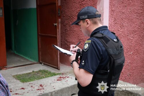 Ukrainian police officer in uniform with a vest writes on a clipboard outside a building; bloodstains on the ground.