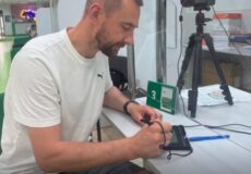 Man in a white t-shirt signs a device at a service desk, with a camera on a tripod nearby in the background.