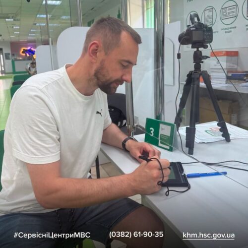 Man in a white t-shirt signs a device at a service desk, with a camera on a tripod nearby in the background.