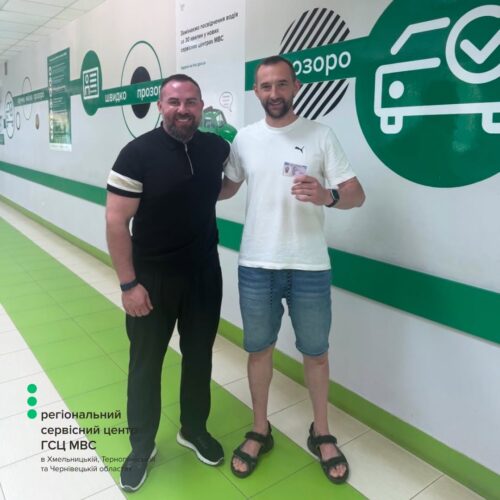 Two men stand in a lime-green, white corridor of a regional service center, smiling for the camera; one in black polo, the other in a white T-shirt and shorts, holding a card.