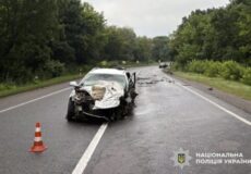 White sedan badly damaged in a road collision on a wet highway, orange traffic cone in front; National Police of Ukraine watermark in corner.