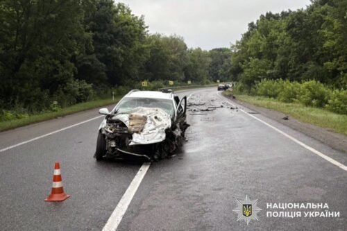 White sedan badly damaged in a road collision on a wet highway, orange traffic cone in front; National Police of Ukraine watermark in corner.