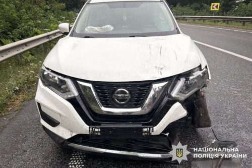 White Nissan with severe front-end damage on a highway, bumper and hood crumpled, left headlight damaged, and police watermark visible in the corner.
