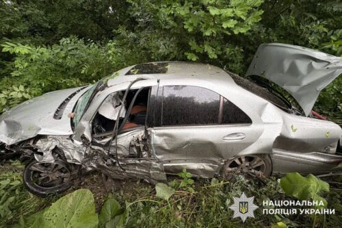 Severely damaged silver sedan after a crash in a leafy roadside area, with the front and side crushed; Ukrainian National Police logo visible in the lower-right corner.
