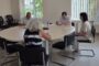 Four adults seated around a round conference table in a bright meeting room; a woman with a striped top is seen from behind while others take notes.
