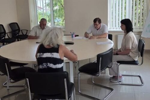 Four adults seated around a round conference table in a bright meeting room; a woman with a striped top is seen from behind while others take notes.