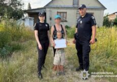 Two Ukrainian police officers stand with a woman and a child holding a document in a grassy yard outside a house.