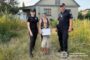Two Ukrainian police officers stand with a woman and a child holding a document in a grassy yard outside a house.