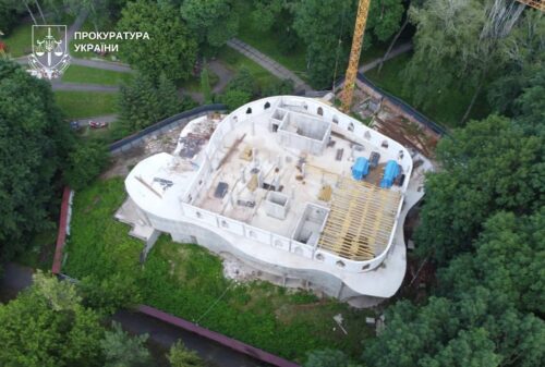 Partially built white concrete structure in a green forested area with a crane overhead; Prosecutor's Office of Ukraine logo in the top-left corner.