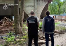 Two men in dark vests stand with their backs to the camera at a construction site beneath a bridge; Ukrainian Prosecutor's Office logo visible in the upper left.