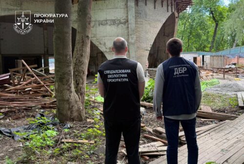 Two men in dark vests stand with their backs to the camera at a construction site beneath a bridge; Ukrainian Prosecutor's Office logo visible in the upper left.