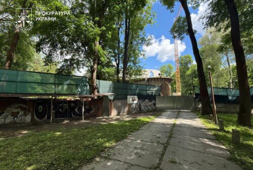 Path through a park toward a construction site behind a green fence; graffiti on the wall, tall trees surround, blue sky visible; Ukrainian Prosecutor's Office watermark in the upper-left corner.