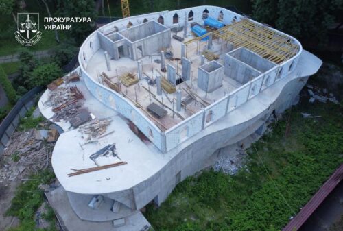 Aerial view of a curved concrete building under construction on a hillside, with exposed framing and formwork; Ukrainian Prosecutor's Office logo in the upper left.