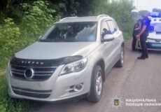 Silver SUV on a rural road with bushes; two police officers stand nearby, Ukrainian National Police emblem in the bottom-right.