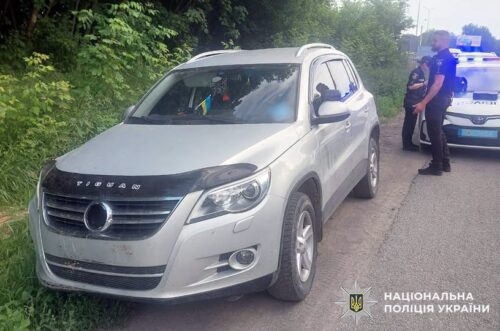 Silver SUV on a rural road with bushes; two police officers stand nearby, Ukrainian National Police emblem in the bottom-right.