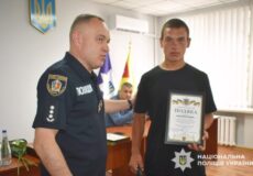 Ukrainian police officer presents a certificate of appreciation to a young man in an office, with the Ukrainian flag and police emblem in view.