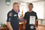 Ukrainian police officer presents a certificate of appreciation to a young man in an office, with the Ukrainian flag and police emblem in view.