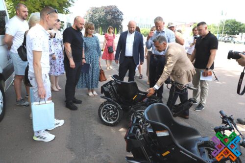 Man in a beige jacket demonstrates a black three‑wheeled motorcycle to a crowd gathered outdoors, as onlookers watch attentively.
