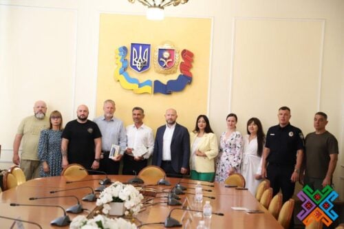 Eleven people stand along a conference table in a meeting room, posing for a photo; a yellow wall panel with a blue-and-yellow coat-of-arms crest is behind them.