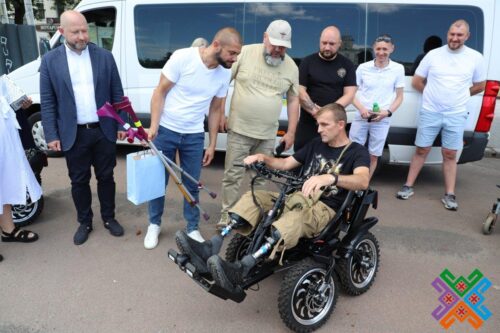 Group of men gathered around a man in a motorized wheelchair outdoors near a white van.