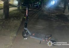 Night scene of a gray electric scooter parked on a sidewalk, stand down, under trees; watermark reads National Police of Ukraine on the bottom right.