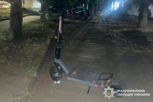 Night scene of a gray electric scooter parked on a sidewalk, stand down, under trees; watermark reads National Police of Ukraine on the bottom right.