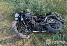 Black motorcycle lying on its side in tall grass, front wheel toward the camera; Ukrainian police watermark in the bottom right corner.