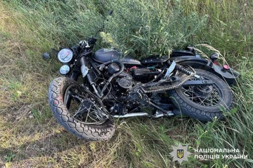 Black motorcycle lying on its side in tall grass, front wheel toward the camera; Ukrainian police watermark in the bottom right corner.