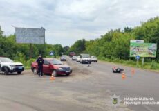 Police at a two-vehicle crash on a rural road: a red car with cones around it, a motorcycle on its side ahead, several bystanders and vehicles in the distance.