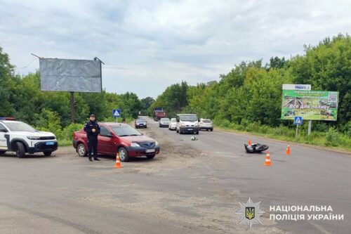 Police at a two-vehicle crash on a rural road: a red car with cones around it, a motorcycle on its side ahead, several bystanders and vehicles in the distance.