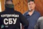 Back view of a man in a black jacket labeled 'контррозвідка СБУ' (SBU counterintelligence) standing in a wooden room with another man whose face is blurred.