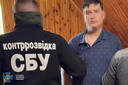 Back view of a man in a black jacket labeled 'контррозвідка СБУ' (SBU counterintelligence) standing in a wooden room with another man whose face is blurred.