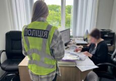 Ukrainian police officer in a bright reflective vest (Poli̇tīia) stands by a desk, reviewing documents as a person sits at the desk in an office setting.