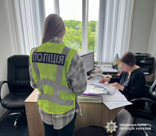 Ukrainian police officer in a bright reflective vest (Poli̇tīia) stands by a desk, reviewing documents as a person sits at the desk in an office setting.