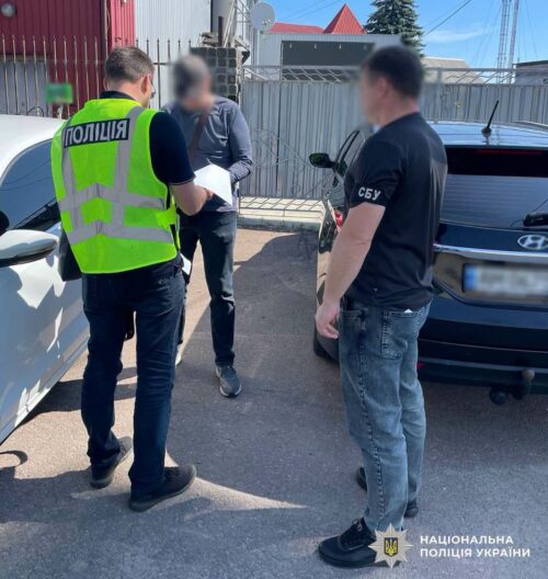 Police officers question a man beside a white car in a parking lot, one in a neon vest and the other in a black shirt; Ukrainian police logo bottom right.