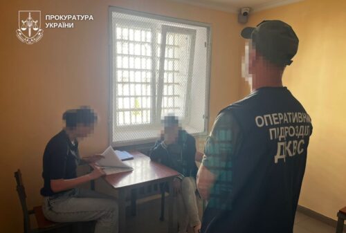 Two men sit at a table with documents in a small yellow room while a third man in a vest stands nearby; a barred window is in the background.
