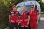 Three emergency responders in red uniforms stand in front of an ambulance on a sunny day, with trees in the background.