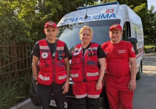 Three emergency responders in red uniforms stand in front of an ambulance on a sunny day, with trees in the background.