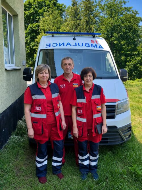 Three paramedics in red uniforms with reflective stripes stand in front of a white ambulance outdoors on a sunny day, with '103' on their shirts and a green building nearby.