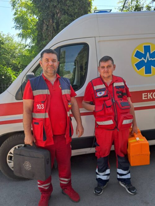 Two paramedics in red uniforms stand beside a white ambulance with a blue Star of Life symbol on a sunny day, one holding a black medical case and the other an orange medical box.