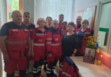 Group of emergency responders in red uniforms posing in a bright room beside a memorial table with a framed photo and flowers.