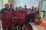 Group of emergency responders in red uniforms posing in a bright room beside a memorial table with a framed photo and flowers.